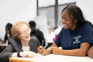 A smiling black student works at her desk as she receives individual support from a teacher.
