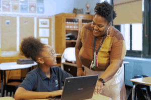 A smiling black female teacher stands next to a young black student working on a laptop at her desk