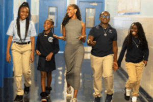 A young female black teacher walks down a hallway, laughing with four young black students